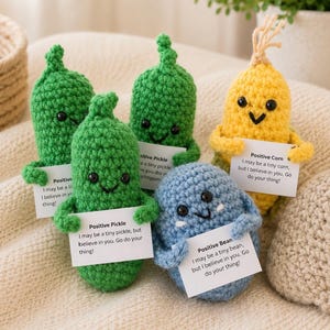 May include: Five small crocheted figures including three green pickles, one yellow corn, and one blue bean. Each figure has black bead eyes, a stitched smile, and holds a white card with text that reads Positive Pickle, Positive Corn, or Positive Bean.