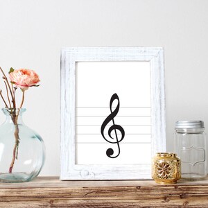 May include: A white framed print with a black treble clef on a white background with five horizontal lines. The print is sitting on a wooden shelf with a clear glass vase with a pink flower and a gold candle holder.