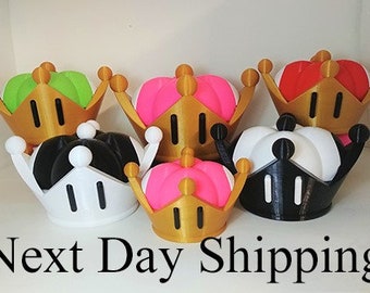 May include: A set of six 3D printed crowns in various colors, including black, white, pink, green, and red. The crowns are all in the same style, with a gold base and a pointed top.