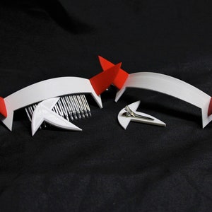 May include: White headband with red devil horns and a comb attachment. The headband is made of plastic and has a simple design.