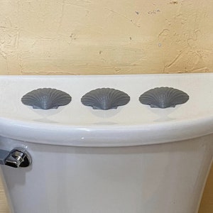 May include: Three grey seashell-shaped toilet tank lid covers on a white toilet tank.
