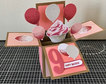 May include: A handmade birthday pop-up card with a cupcake and balloons. The card is pink, red, and silver with the number 9 and the words "Happy Birthday". The card is made of paper and has a 3D effect.