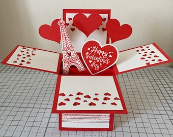 May include: A red and white pop-up Valentine's Day card featuring a paper Eiffel Tower and heart cutouts. The card has a heart-shaped message that says "Happy Valentine's Day".