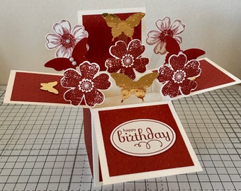 May include: A handmade birthday pop-up card with a red and cream color scheme. The card features red flowers, gold butterflies, and the words "happy birthday" on the front. The card is open, revealing the 3D floral and butterfly design.