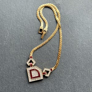 May include: Gold-toned necklace with a geometric pendant. The pendant features a square design with a red center and clear crystal accents. The necklace has a flat chain and a clasp closure.