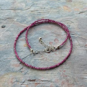 May include: A delicate necklace with small, faceted, deep pink gemstones. The necklace is strung on a fine chain with a silver-coloured clasp and extender. The gemstones are a vibrant shade of pink, creating a subtle sparkle.