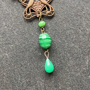 Peut inclure: Une perle de verre verte et blanche ray&eacute;e pend d'une cha&icirc;ne en laiton avec une perle de verre en forme de larme verte au bas. La cha&icirc;ne est attach&eacute;e &agrave; une pi&egrave;ce d&eacute;corative en laiton avec un motif floral.