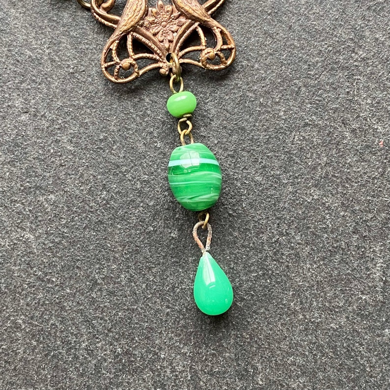 Peut inclure: Une perle de verre verte et blanche ray&eacute;e pend d'une cha&icirc;ne en laiton avec une perle de verre en forme de larme verte au bas. La cha&icirc;ne est attach&eacute;e &agrave; une pi&egrave;ce d&eacute;corative en laiton avec un motif floral.
