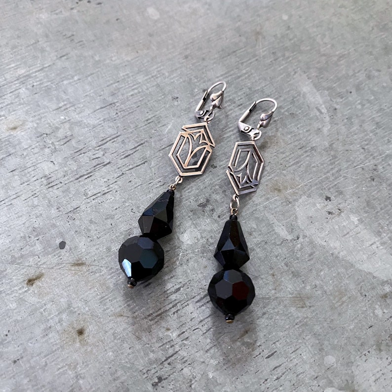 May include: A pair of long silver earrings with black faceted glass beads. The earrings feature a geometric design with a floral motif.