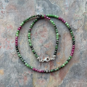 Op de afbeelding: Een ketting bestaande uit kleine, ronde kralen in groen, zwart en magenta. De kralen zijn aan elkaar geregen en vormen een cirkel met een zilveren sluiting. De ketting wordt gepresenteerd op een getextureerd, grijs oppervlak.