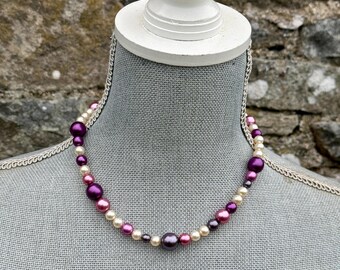 May include: A beaded necklace featuring a mix of purple, pink, and cream-colored faux pearls. The necklace is displayed on a gray mannequin bust against a stone wall background. The pearls vary in size and shade, creating a classic and elegant look.