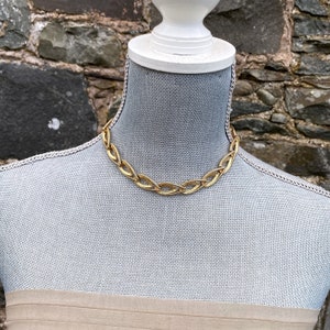 May include: A gold chain necklace with a unique, interwoven design. The necklace is displayed on a mannequin.