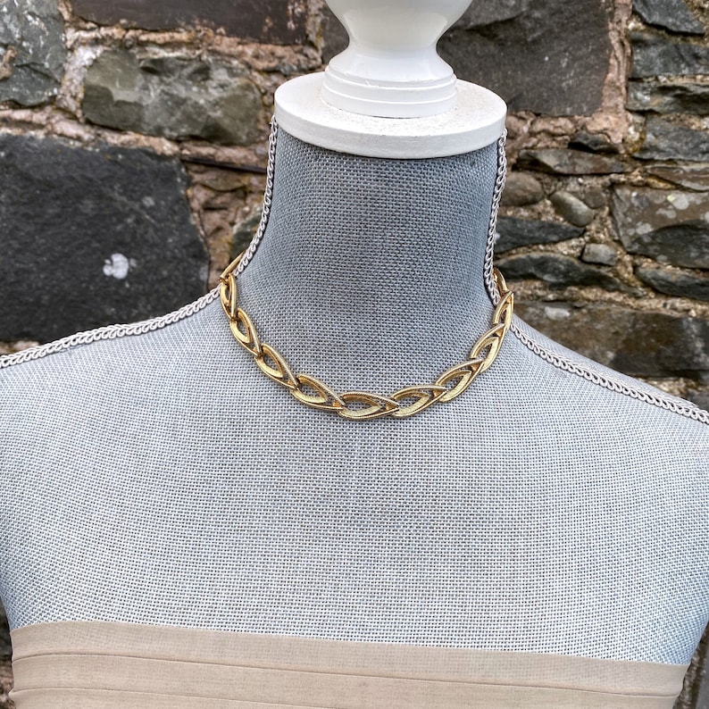 May include: A gold chain necklace with a unique, interwoven design. The necklace is displayed on a mannequin.
