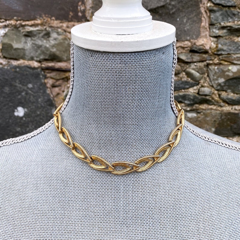 May include: A gold chain necklace with a unique, interlocking design. The necklace is made of thick, chunky links and has a modern, geometric look.