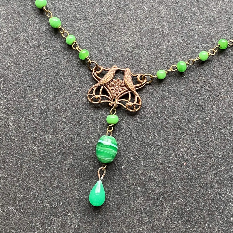 Peut inclure: Un collier de perles de verre vert avec un pendentif en bronze repr&eacute;sentant deux oiseaux et un motif floral. Le pendentif est attach&eacute; &agrave; une perle de verre verte et &agrave; une perle de verre verte en forme de larme.
