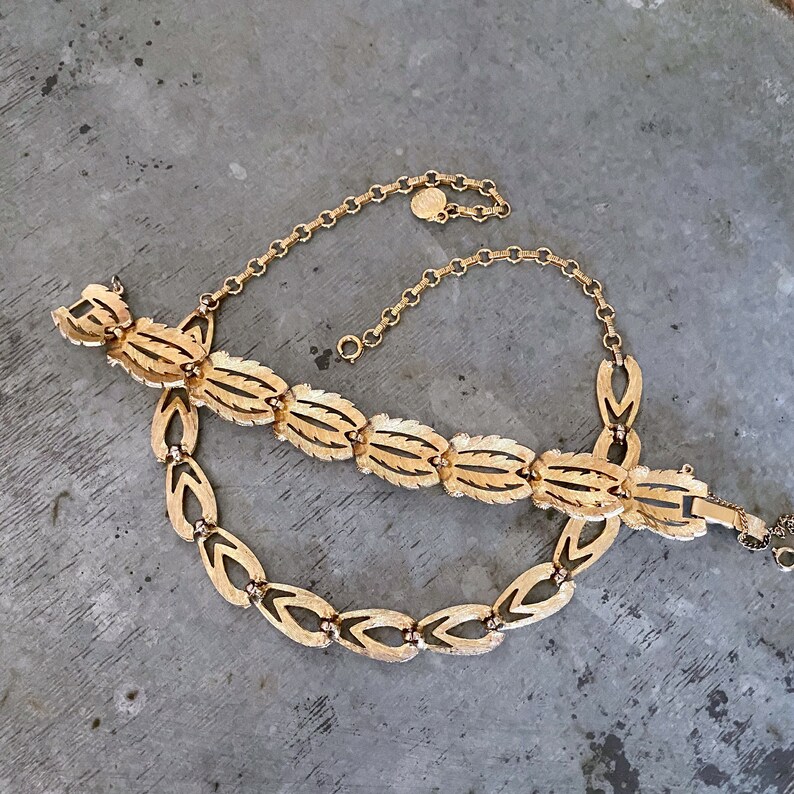 May include: A gold-tone necklace with a delicate chain and a pendant featuring a leaf design. The necklace is made of metal and has a clasp closure.