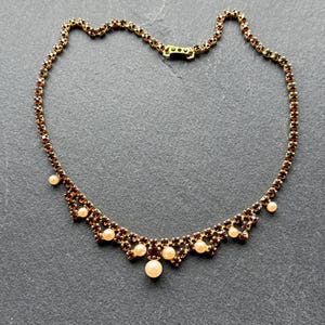 May include: A gold-toned necklace with a series of small, dark red gemstones and cream-colored pearls. The necklace has a decorative, scalloped design at the front and a clasp closure. The necklace is displayed on a dark gray surface.