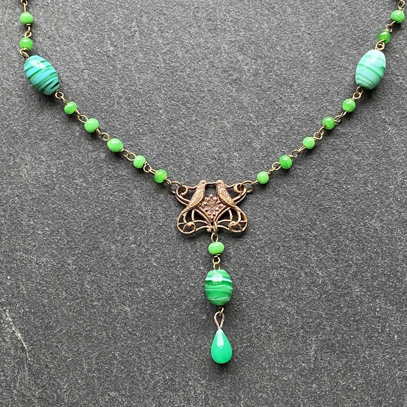 Peut inclure: Un collier de perles de verre vert avec un pendentif en or repr&eacute;sentant deux oiseaux. Le pendentif est attach&eacute; &agrave; une perle de verre verte.