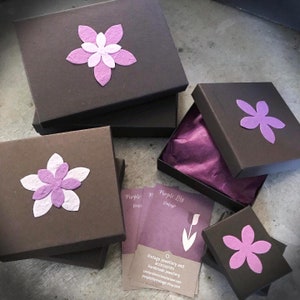 May include: Three brown gift boxes with purple paper flowers on top. The boxes are stacked on top of each other. The top box is open and has a purple tissue paper inside. There is a business card on the table that says "Purple Lily Vintage" and has the text "Vintage jewellery and accessories Handmade jewellery contact@ecochnctalane.com purplelilyvintage.etsy.com".