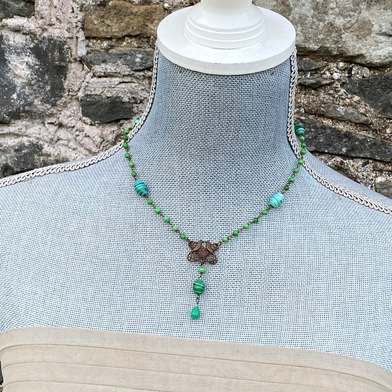 Peut inclure: Un collier vert et bronze avec un pendentif d&eacute;coratif en forme de papillon. Le collier est compos&eacute; de petites perles rondes et d'une perle en forme de larme &agrave; l'extr&eacute;mit&eacute;.