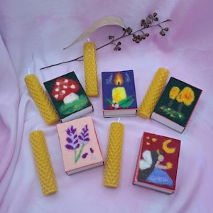 May include: A collection of handmade candles and decorative matchboxes. The candles are yellow and cylindrical. The matchboxes feature felted designs of a mushroom, a candle, lavender, and a fairy. The background is a soft pink fabric.