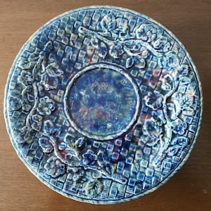 May include: A blue and white ceramic plate with a raised design of flowers and leaves. The plate has a textured surface and a raised rim.