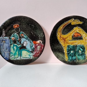 May include: Two ceramic plates with colourful designs. One plate features a black background with a scene of a person in a green and blue robe with a white animal. The other plate features a black background with a yellow camel with a green and blue house.