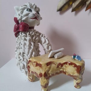 May include: A ceramic cat figurine seated at a small table. The cat is white with a red bow and a serious expression. The table is tan with brown accents, and there are small objects on top. The figurine is likely hand-painted.