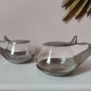 May include: Two vintage glass and metal sugar bowls. Each bowl has a clear glass body with a rounded shape and a metal base, handle, and lid. The lids have a small, curved handle. The bowls are displayed on a white surface.