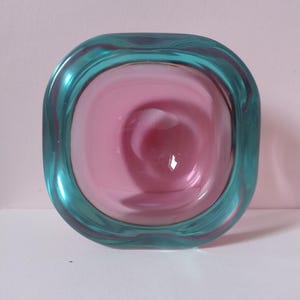 May include: A square glass bowl with rounded corners. The bowl has a teal rim and a pink and white swirled interior. The glass appears transparent. The bowl is likely for decorative purposes. The measurements are in centimetres.