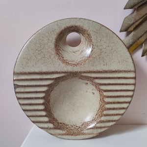 May include: A round, ceramic decorative plate in beige and brown. It has a circular indentation with a hole at the top and horizontal lines at the bottom. The plate's surface is textured. The plate is likely for decorative purposes.
