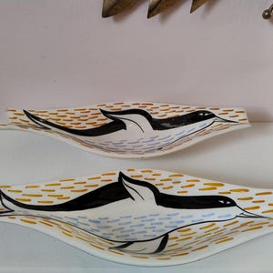 May include: Two white ceramic serving dishes with a bird design. Each dish features a black bird silhouette with a long beak, set against a background of orange and brown dashes. The bird's body has blue and purple accents.