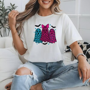 May include: White t-shirt featuring three ghost figures with colorful leopard print and bow details. The ghosts are in teal, pink, and leopard print, with bats and stars. A fun and festive design for Halloween.