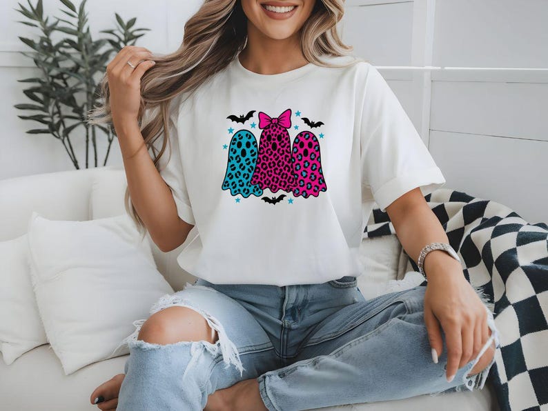 May include: White t-shirt featuring three ghost figures with colorful leopard print and bow details. The ghosts are in teal, pink, and leopard print, with bats and stars. A fun and festive design for Halloween.