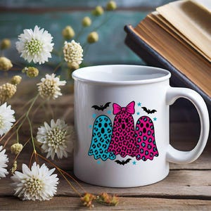 May include: White ceramic mug featuring three ghost illustrations with leopard print patterns in teal, pink, and red. The ghosts are decorated with bats, stars, and a pink bow. The mug is on a wooden surface with flowers and a book.