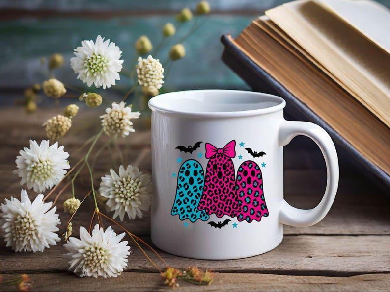 May include: White ceramic mug featuring three ghost illustrations with leopard print patterns in teal, pink, and red. The ghosts are decorated with bats, stars, and a pink bow. The mug is on a wooden surface with flowers and a book.