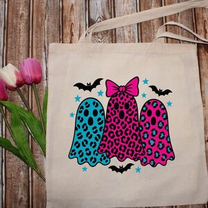 May include: Beige tote bag featuring three ghost illustrations with leopard print patterns in blue and pink. The ghosts are decorated with black bats and blue stars, perfect for Halloween.