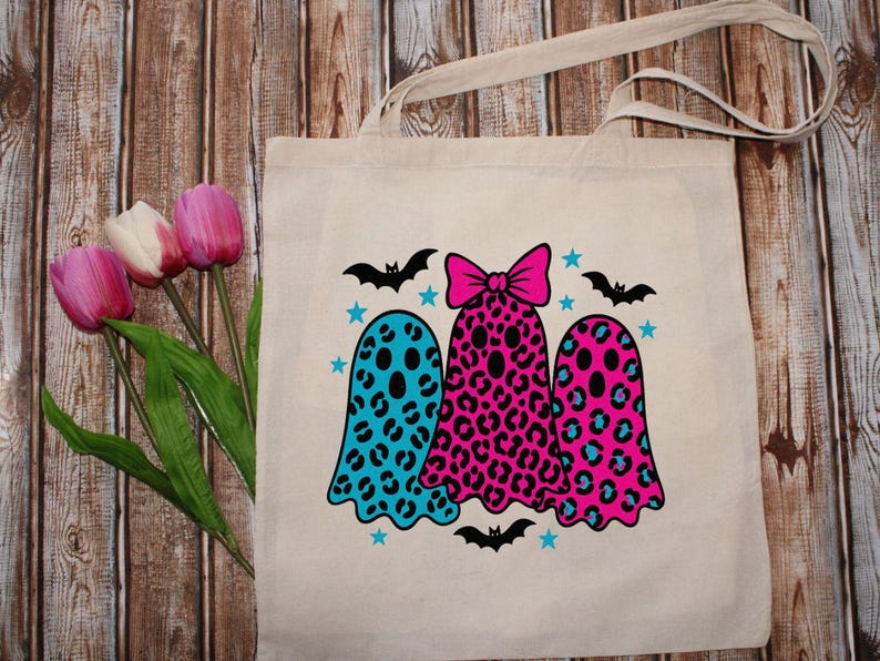 May include: Beige tote bag featuring three ghost illustrations with leopard print patterns in blue and pink. The ghosts are decorated with black bats and blue stars, perfect for Halloween.