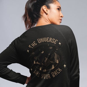 May include: Black long-sleeve sweatshirt with a gold graphic print of a mountain range, stars, and a crescent moon. The text "The Universe Has Your Back" is printed in a circle around the graphic.