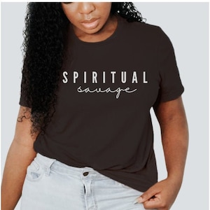 May include: A dark brown t-shirt with the words "SPIRITUAL savage" printed in white. The shirt is a crew neck style and is being worn with light blue jeans.