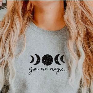 May include: A gray sweatshirt with a black graphic of a crescent moon, full moon, and a crescent moon with stars. The text "you are magic." is printed below the moons.