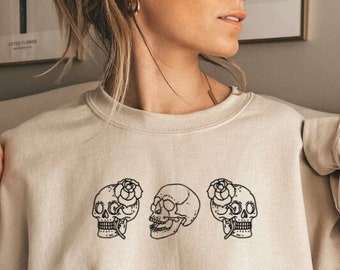 May include: Beige sweatshirt featuring a black line art design of three skulls. Two skulls have a rose on the forehead. The sweatshirt has long sleeves and a crew neck. The design is centered on the front of the shirt.