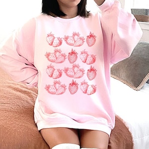 May include: A pink sweatshirt with a graphic print of red strawberries. The sweatshirt has long sleeves and a crew neck.