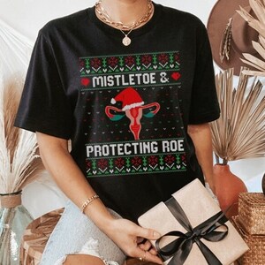 Peut inclure: T-shirt noir avec un motif sur le thème de Noël. Le motif comprend le texte "Mistletoe & Protecting Roe" avec un utérus portant un bonnet de Père Noël. Le t-shirt présente un motif festif en rouge, vert et blanc.