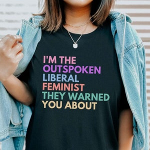 May include: A black t-shirt with the text "I'm the Outspoken Liberal Feminist They Warned You About" in rainbow colors.