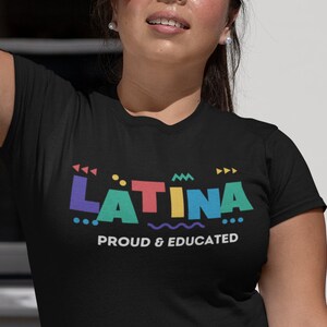 May include: Black t-shirt with the word "LATINA" in colorful geometric letters and the phrase "PROUD & EDUCATED" in white. The model is wearing blue jeans and is shielding her eyes from the sun.