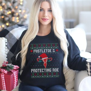 Peut inclure: Sweat-shirt noir avec l'inscription "Mistletoe & Protecting Roe" et un motif festif. Le graphisme comprend un utérus portant un bonnet de Père Noël, entouré de cœurs et de motifs de vacances. Le sweat-shirt est porté avec un jean bleu.