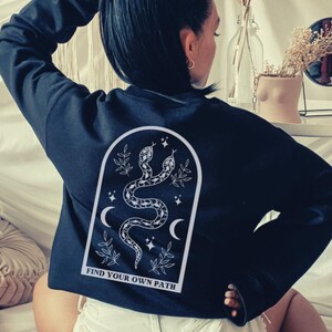 May include: Black sweatshirt with a white graphic on the back featuring two snakes, crescent moons, stars, and the text "FIND YOUR OWN PATH". The sweatshirt is a crew neck style.