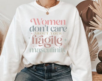 以下が含まれることがあります： 「Women don't care about your fragile masculinity」というピンク、青、薄ピンクの文字がプリントされた白いスウェットシャツ。スウェットシャツはブルージーンズと組み合わせています。