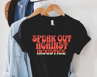 May include: Black t-shirt with the words "SPEAK OUT AGAINST INJUSTICE" in orange, retro-style lettering. The bottom of the text has a green and red stripe design. The shirt is hanging on a wooden hanger.